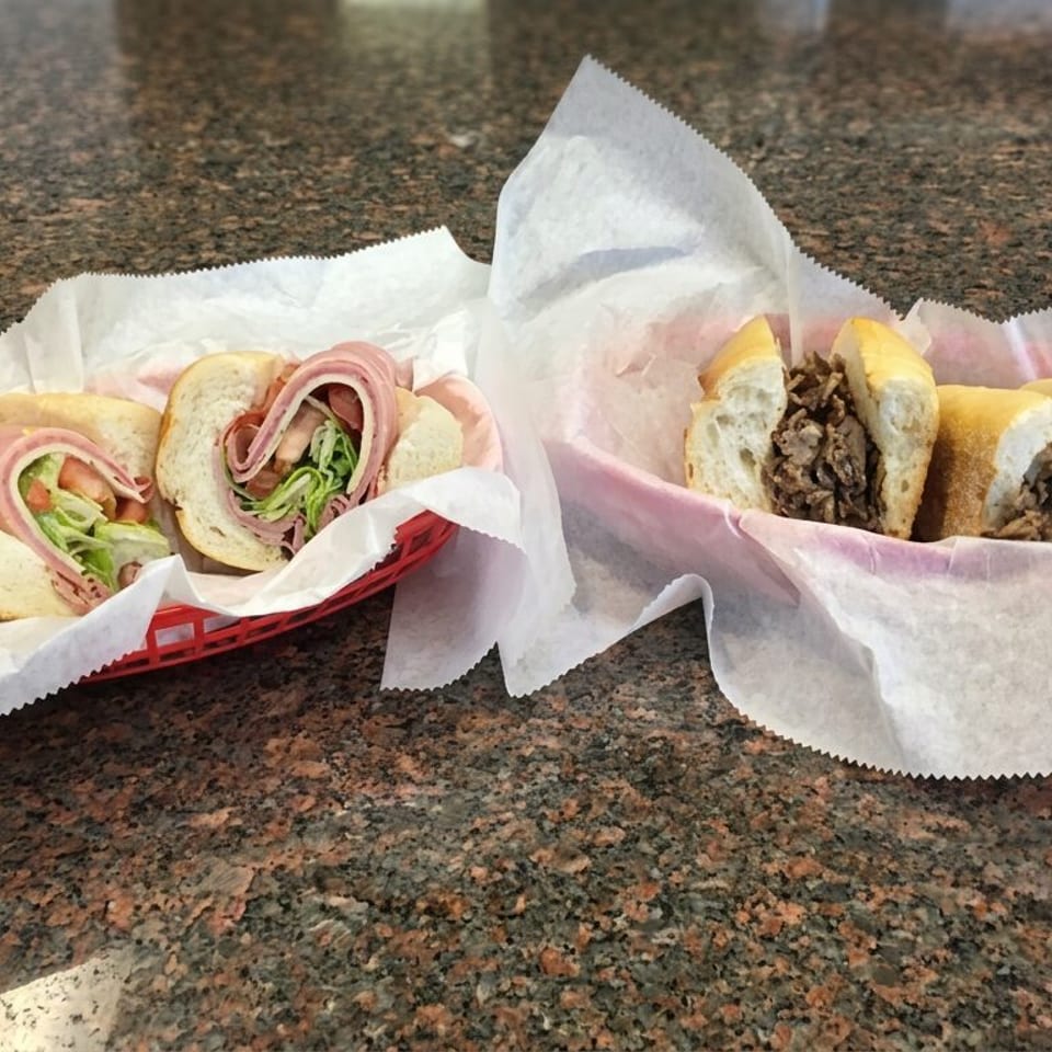 Two baskets hold a sliced deli sandwich with lettuce and a sliced cheesesteak sandwich on a granite countertop.